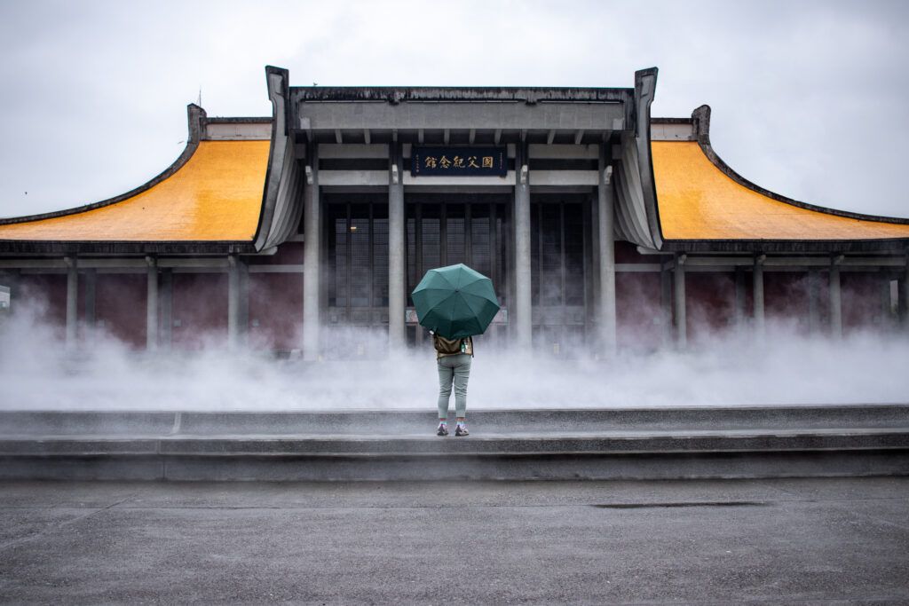 A low-angle shot of the National Dr. Sun Yat-sen Memorial Hall in Taipei, featuring its distinctive yellow streamlined roof and a person with a green umbrella standing in the misty foreground.